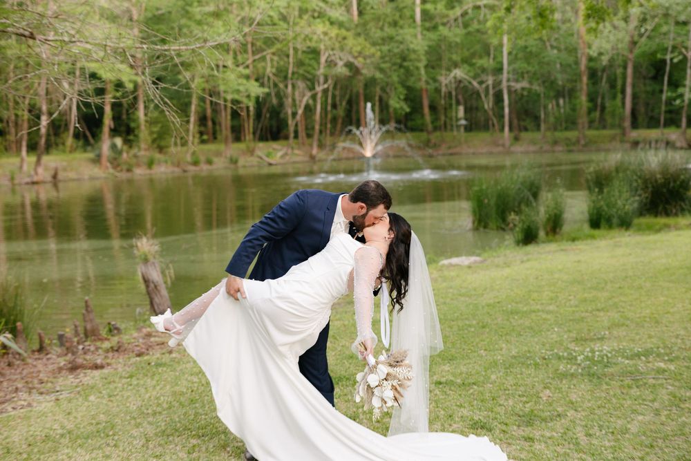 A wedding couple embraces in a sunlit meadow surrounded by lush green trees and a serene lake in the background.
