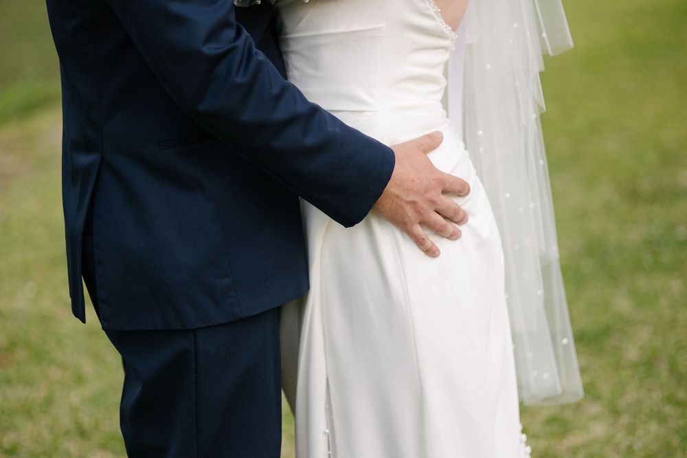 Close-up view of a bride in white dress and groom in navy suit embracing on grassy lawn during wedding ceremony.