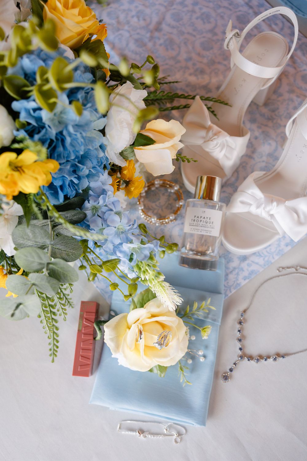 Elegant floral arrangement with yellow roses and blue flowers alongside perfume bottles on a light surface.