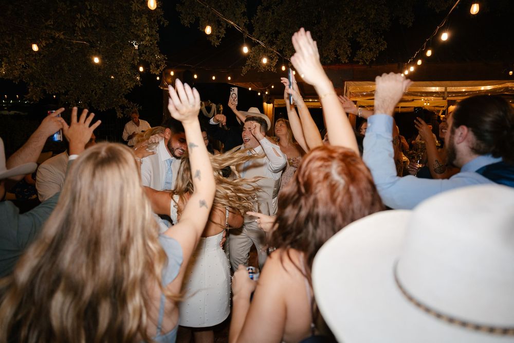 A lively crowd dances with raised hands under string lights at an evening celebration party.