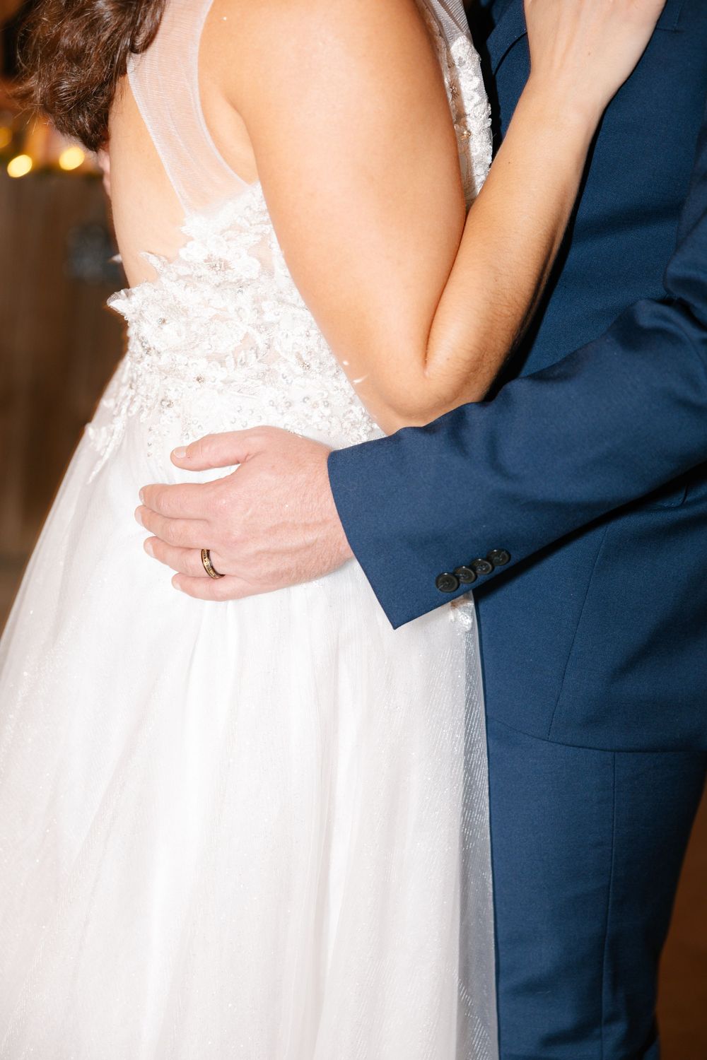 Close-up details of a white lace wedding dress and navy suit during an embrace.
