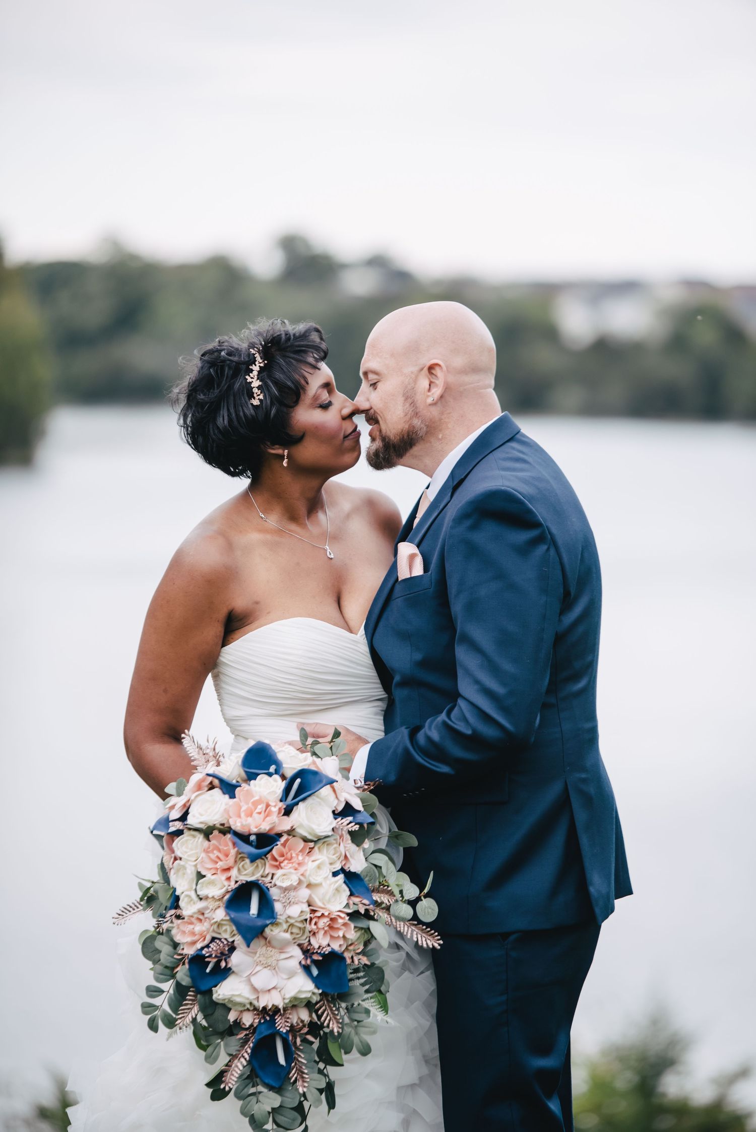 A tender wedding moment captured by a lake with a beautiful floral bouquet in soft tones.