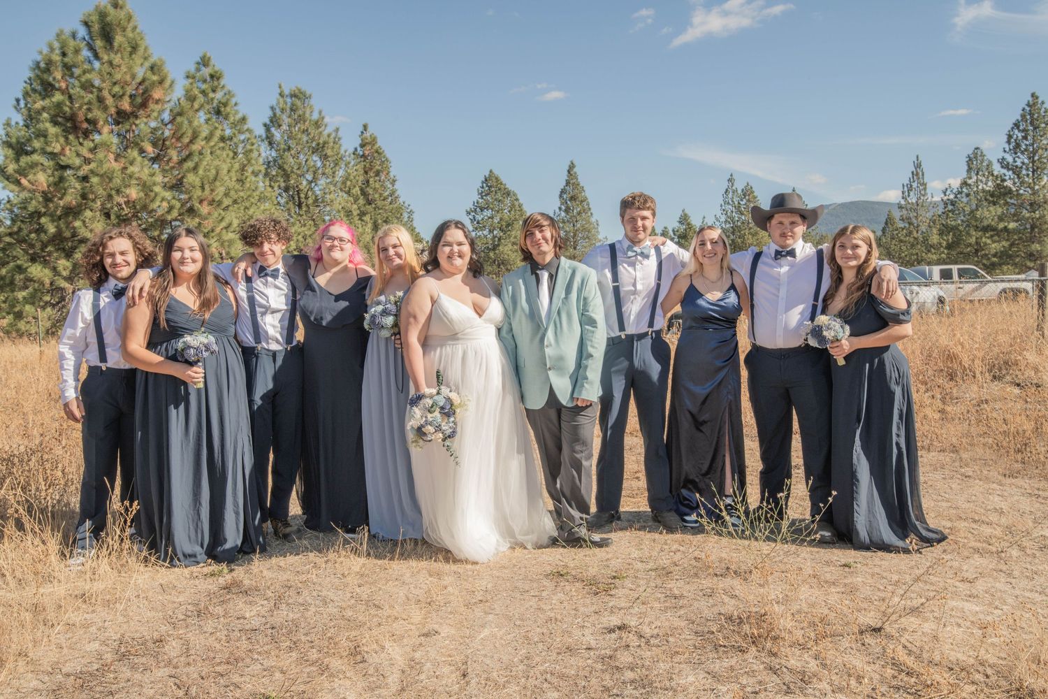 Wedding party posing outdoors in rustic field setting with bridesmaids in blue gowns and groomsmen in suspenders.