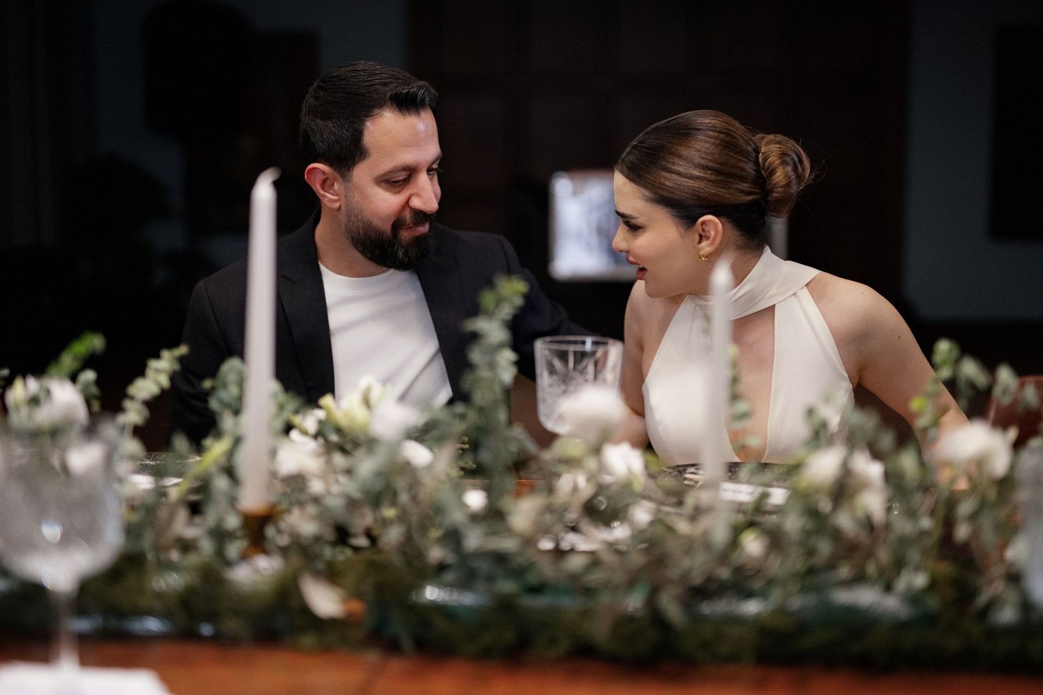 Couple smiling at each other at elegantly decorated wedding reception table with candles and greenery.