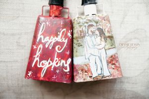 Two decorated cow bells with custom artwork and text displayed on white fabric.