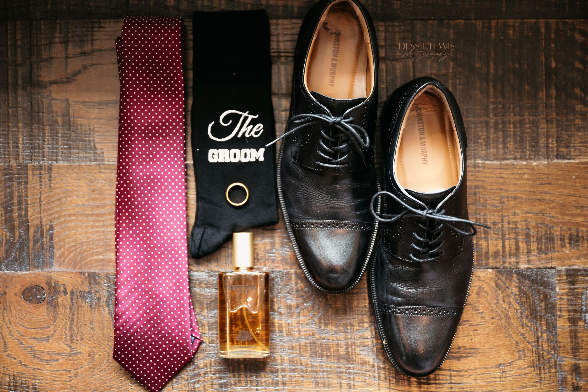 Men's formal accessories including red tie, dress shoes and cologne on wooden surface.