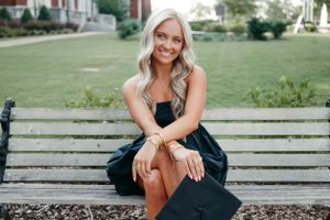 Graduate in a black dress sits casually on a wooden bench in a garden setting.