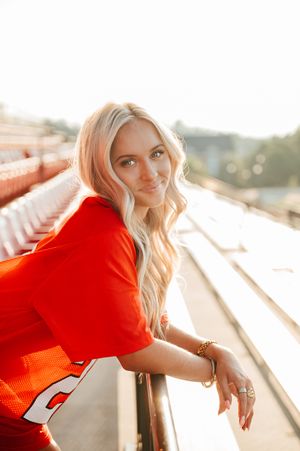 Graduate in football jersey leans against stadium railing during sunset.
