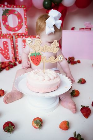 Pink birthday cake on white pedestal with strawberry decor and red balloon backdrop.