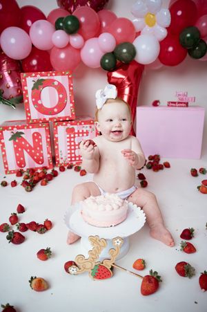 Festive pink and red cake smash scene with letter blocks, balloons, and scattered strawberries.