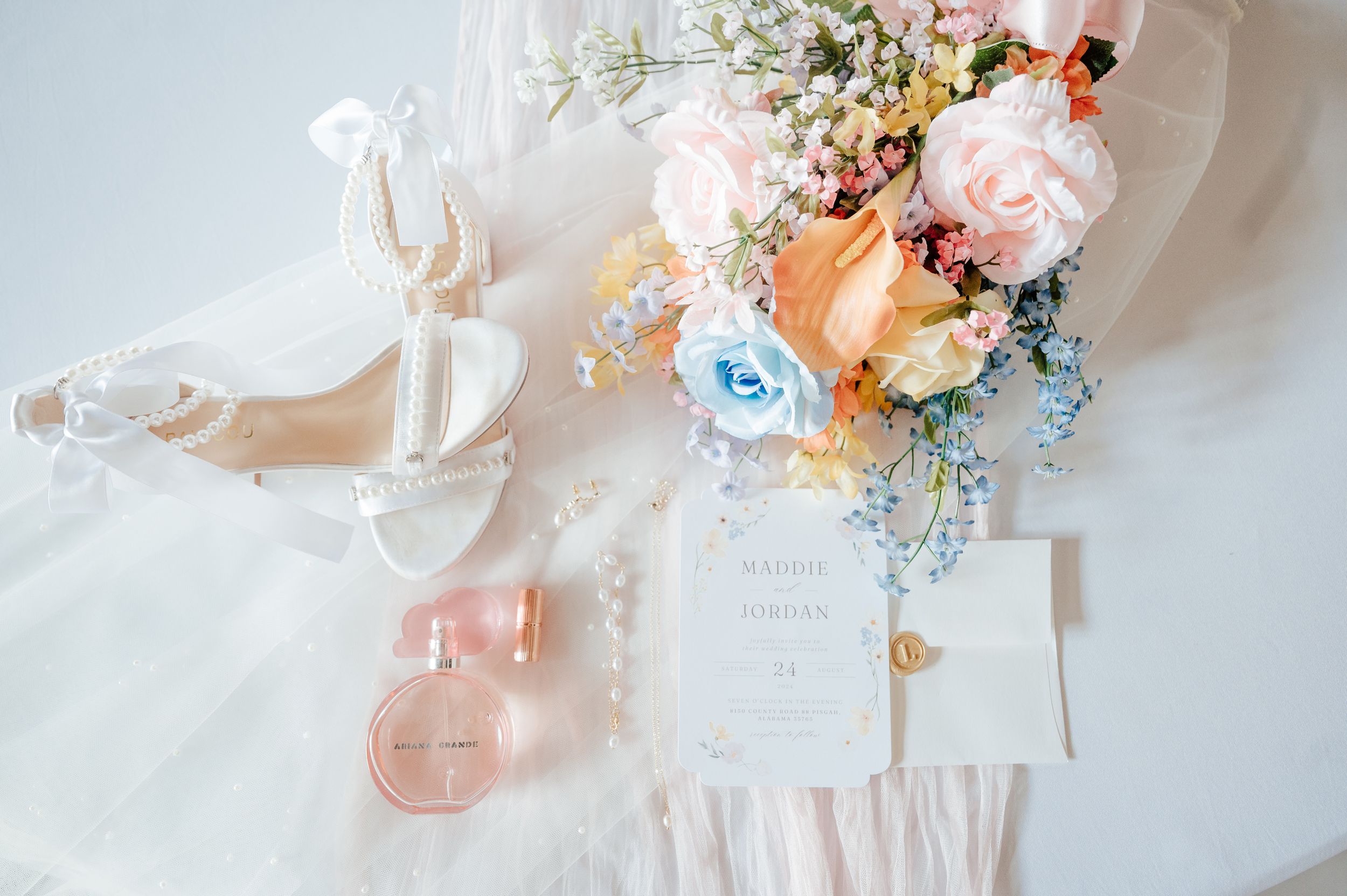 Wedding details flatlay featuring pastel flowers, invitation and bridal shoes on white fabric.