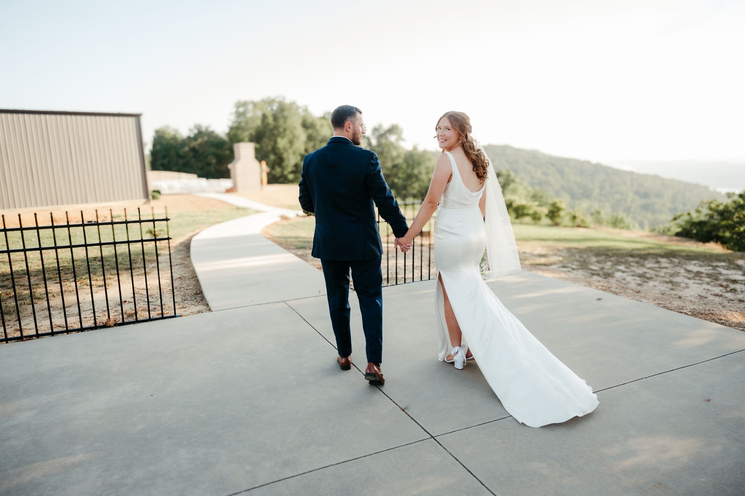 A couple walks hand in hand along an outdoor venue path with scenic mountain views.