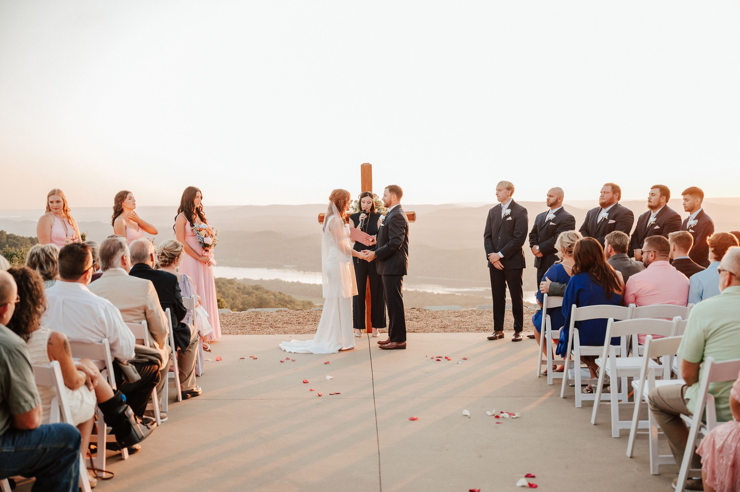 Outdoor wedding ceremony setup with guests seated on white chairs at sunset.