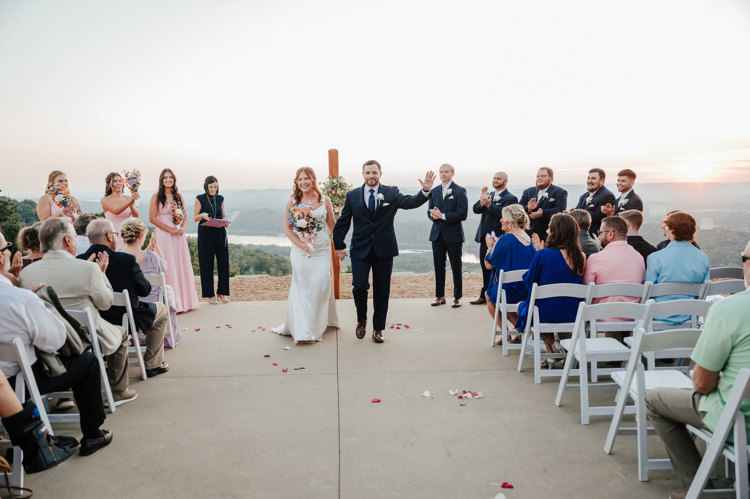 Wedding ceremony procession on an outdoor patio with guests seated on white chairs.