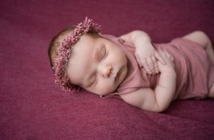 Sleeping newborn baby wearing pink flower crown posed on burgundy fabric.