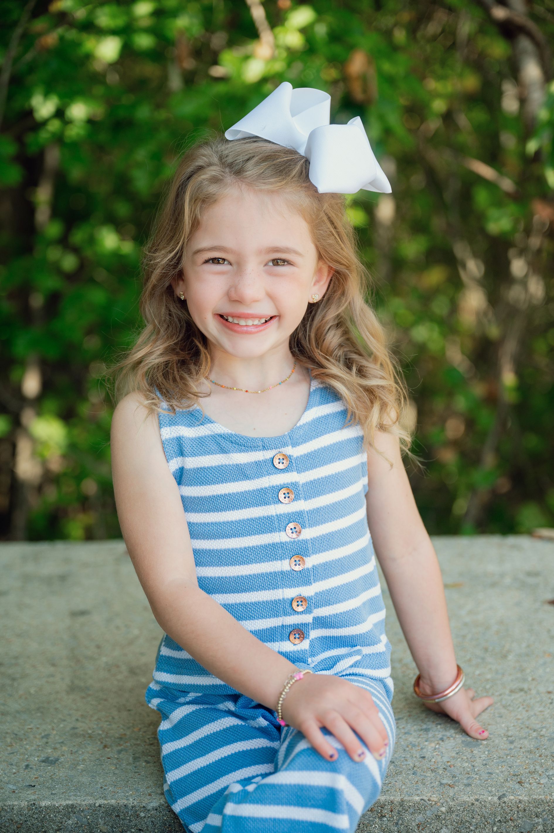 Young person in blue striped outfit and white bow sitting outdoors.