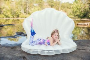 Fantasy-themed photo with mermaid tail costume in large decorative shell prop by pond.