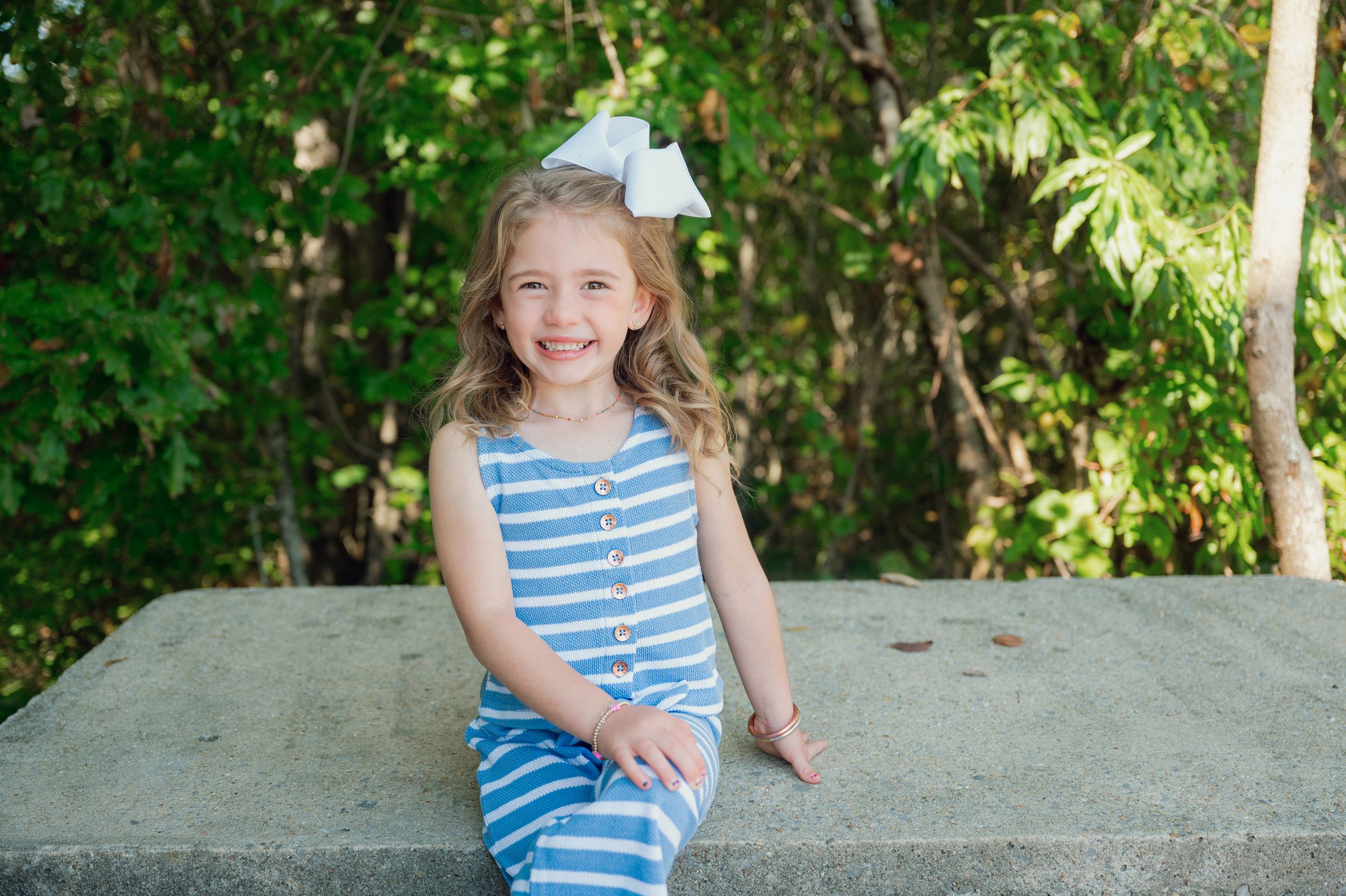 Child wearing blue striped outfit and white bow sitting on concrete surface.