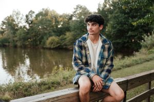 Outdoor portrait of someone wearing plaid and shorts while sitting on wooden bench near water.
