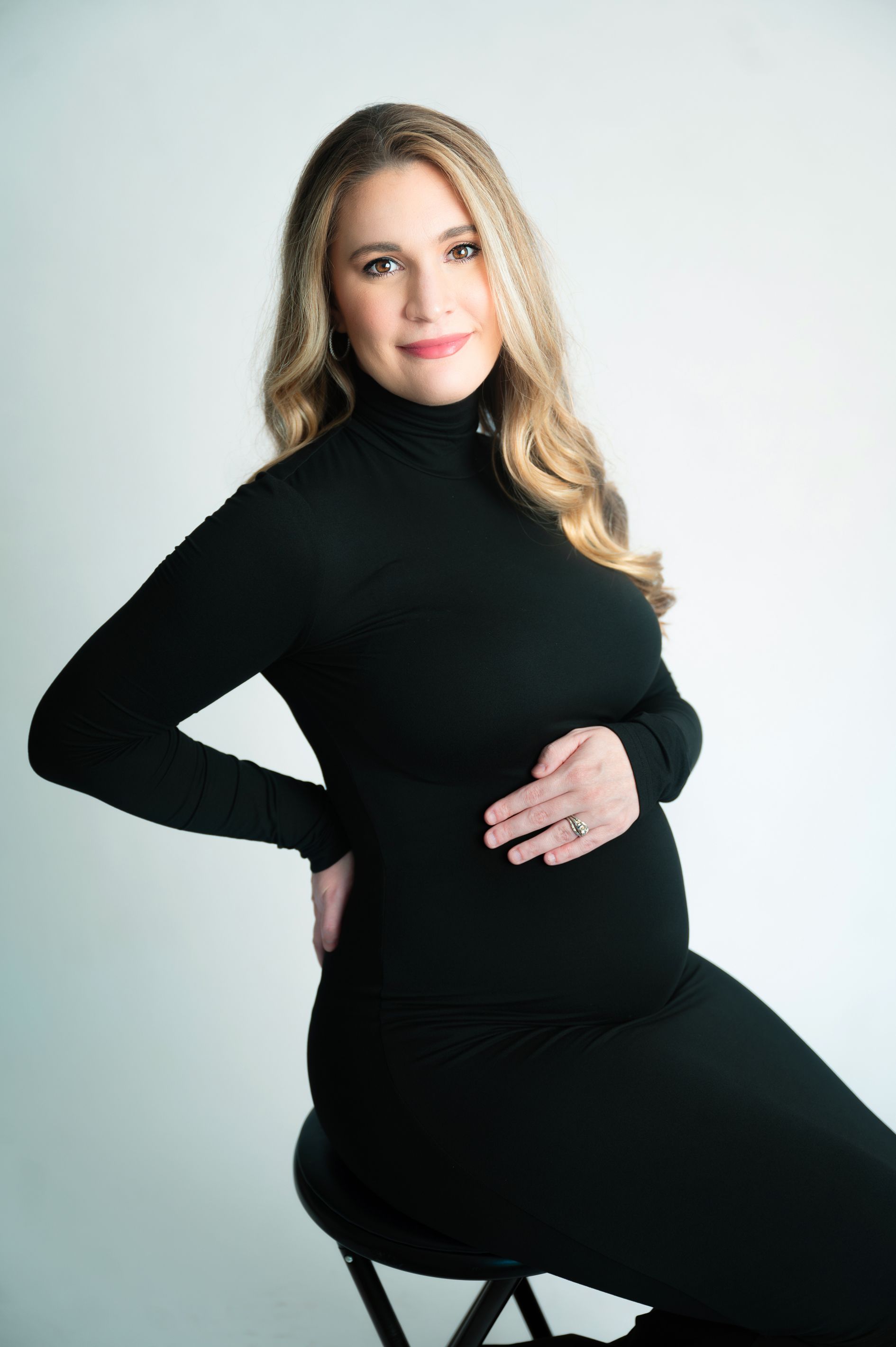 Studio maternity portrait of woman in black turtleneck dress against light background.