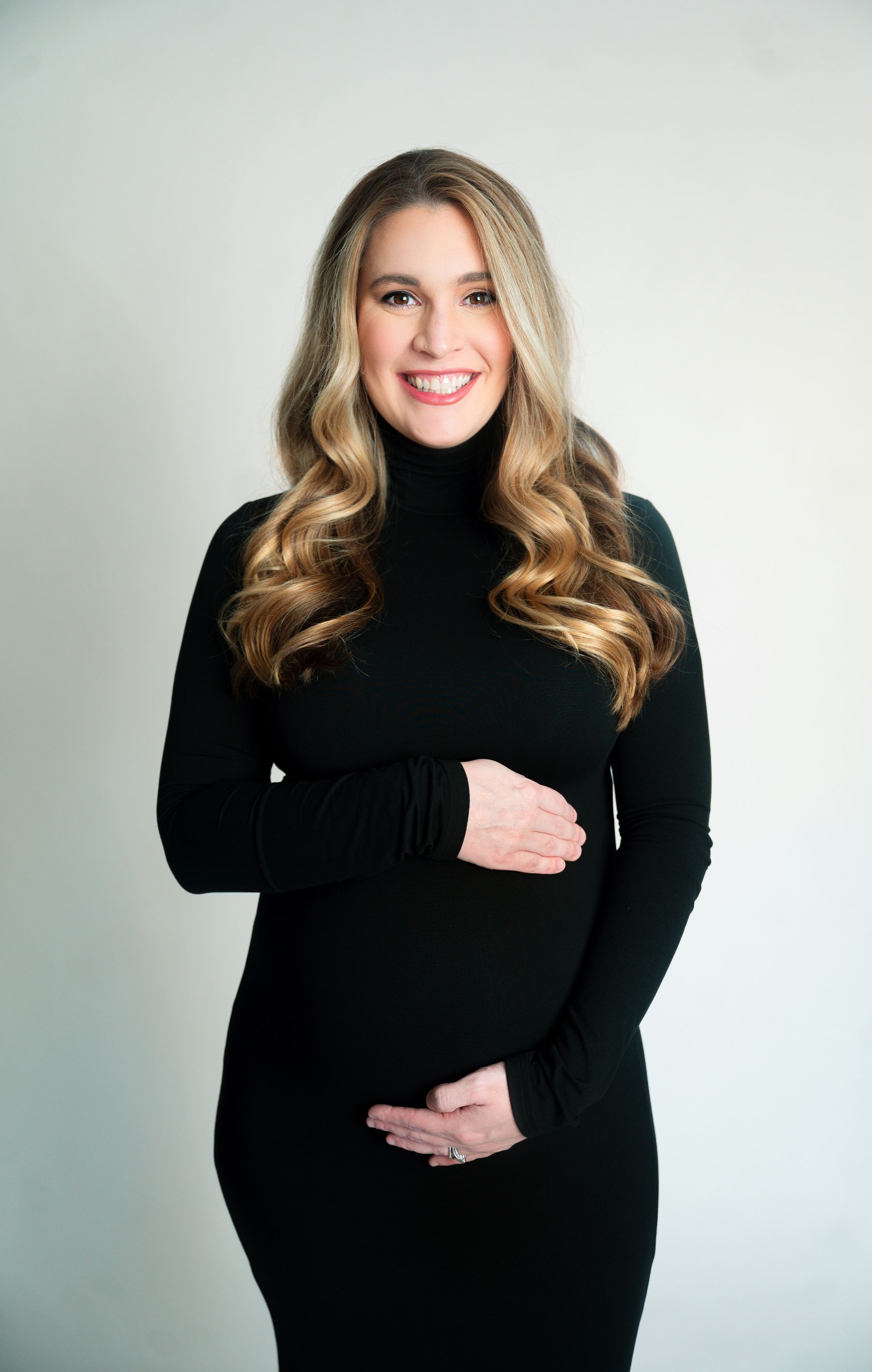 Maternity portrait of woman in black dress smiling against light studio background.