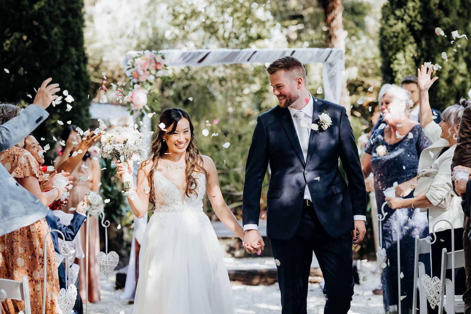 Wedding guests throw confetti as a couple walks down the aisle under natural outdoor lighting.