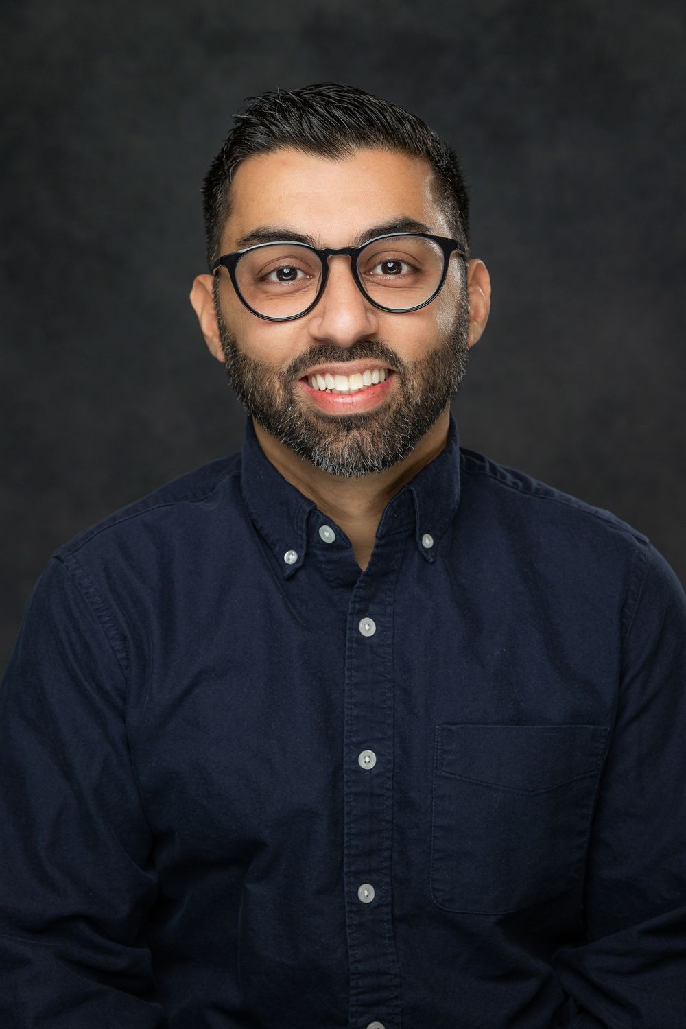 Professional headshot of a person wearing glasses and a navy button-down shirt against a dark background.