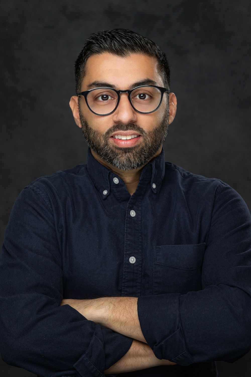 Professional headshot of someone with glasses and a beard wearing a navy button-down shirt against a dark background.