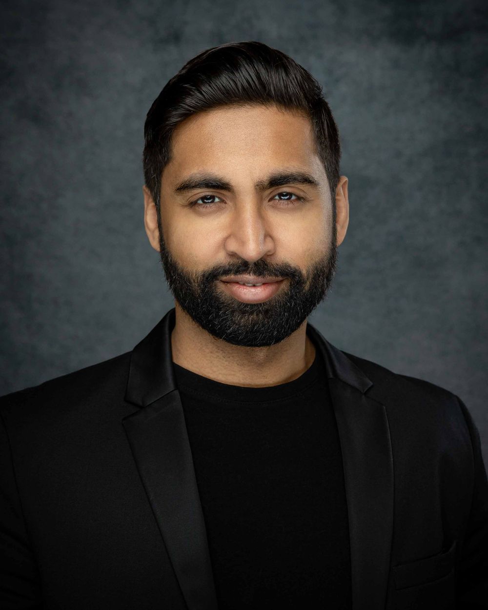 Professional headshot portrait with black blazer and black shirt against grey studio background.