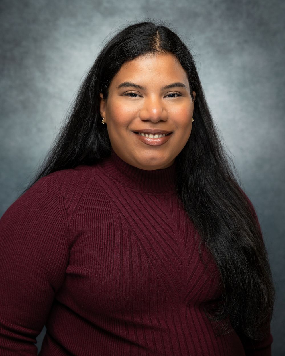 Professional studio portrait of someone with long dark hair wearing a burgundy sweater against a gray background.