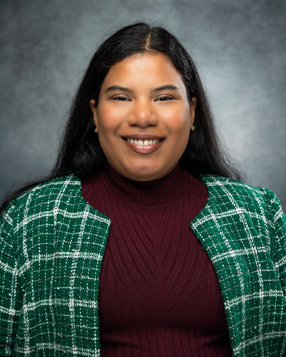 Professional headshot portrait of someone wearing a burgundy turtleneck and green plaid cardigan against a gray background.