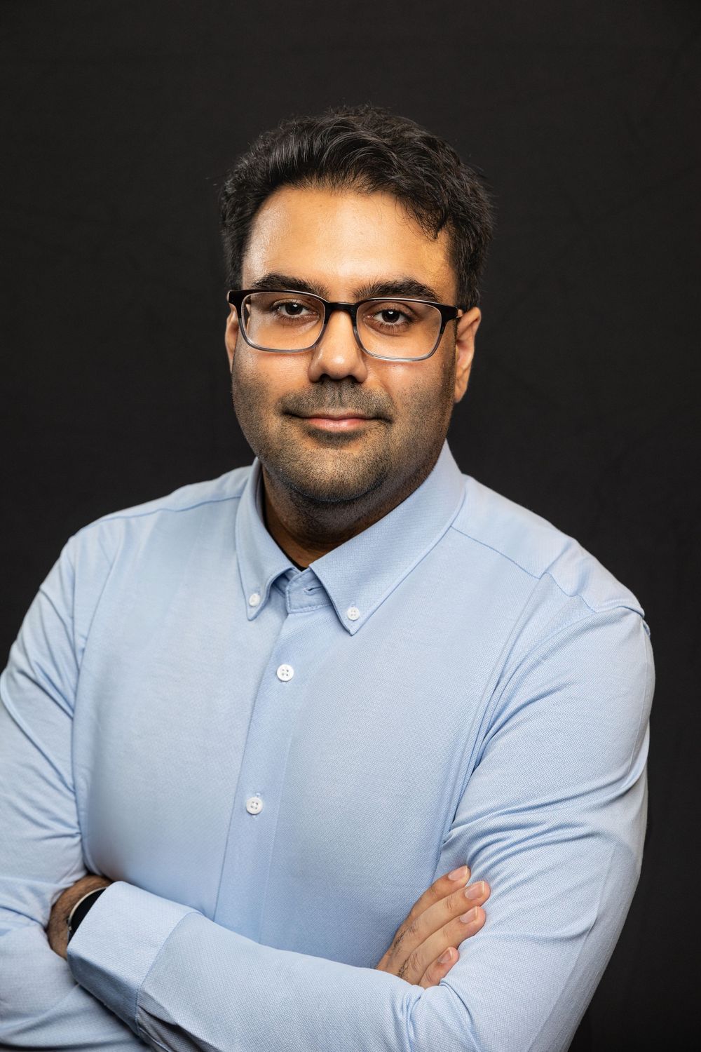 Professional headshot of person in light blue button-down shirt and glasses against dark background with arms crossed.