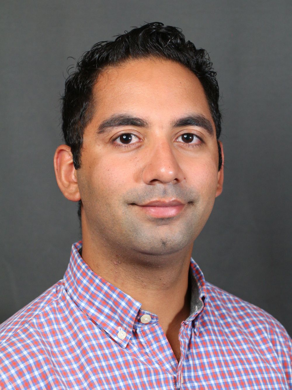 Professional headshot of person in pink and blue checkered button-down shirt against gray background.