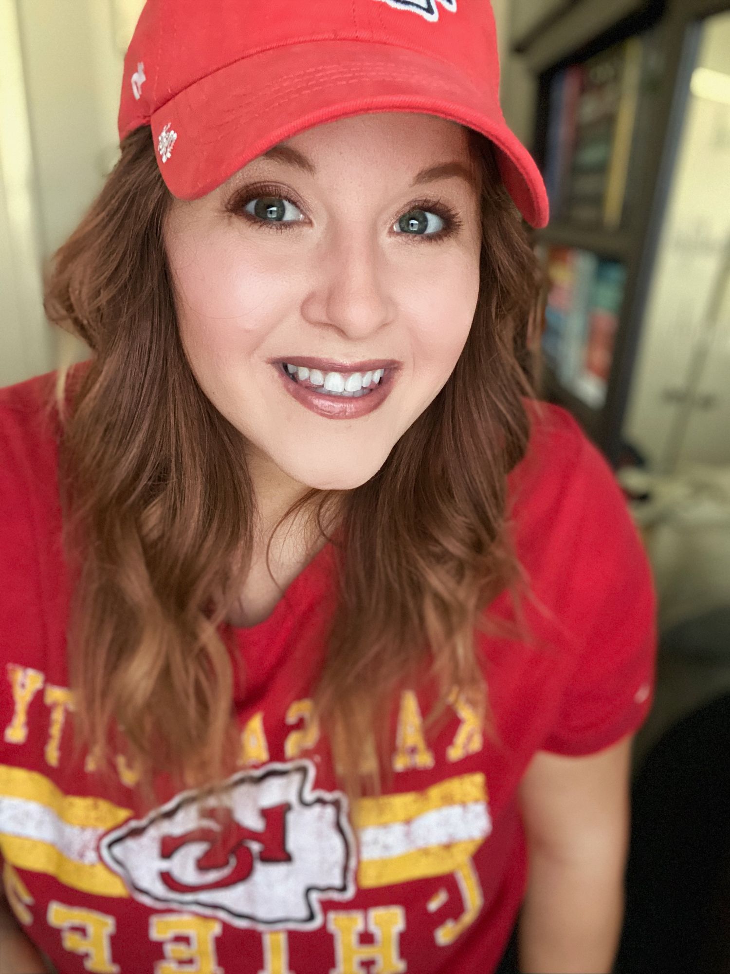 Smiling woman wearing red Kansas City Chiefs cap and jersey taking a selfie at a sports venue.