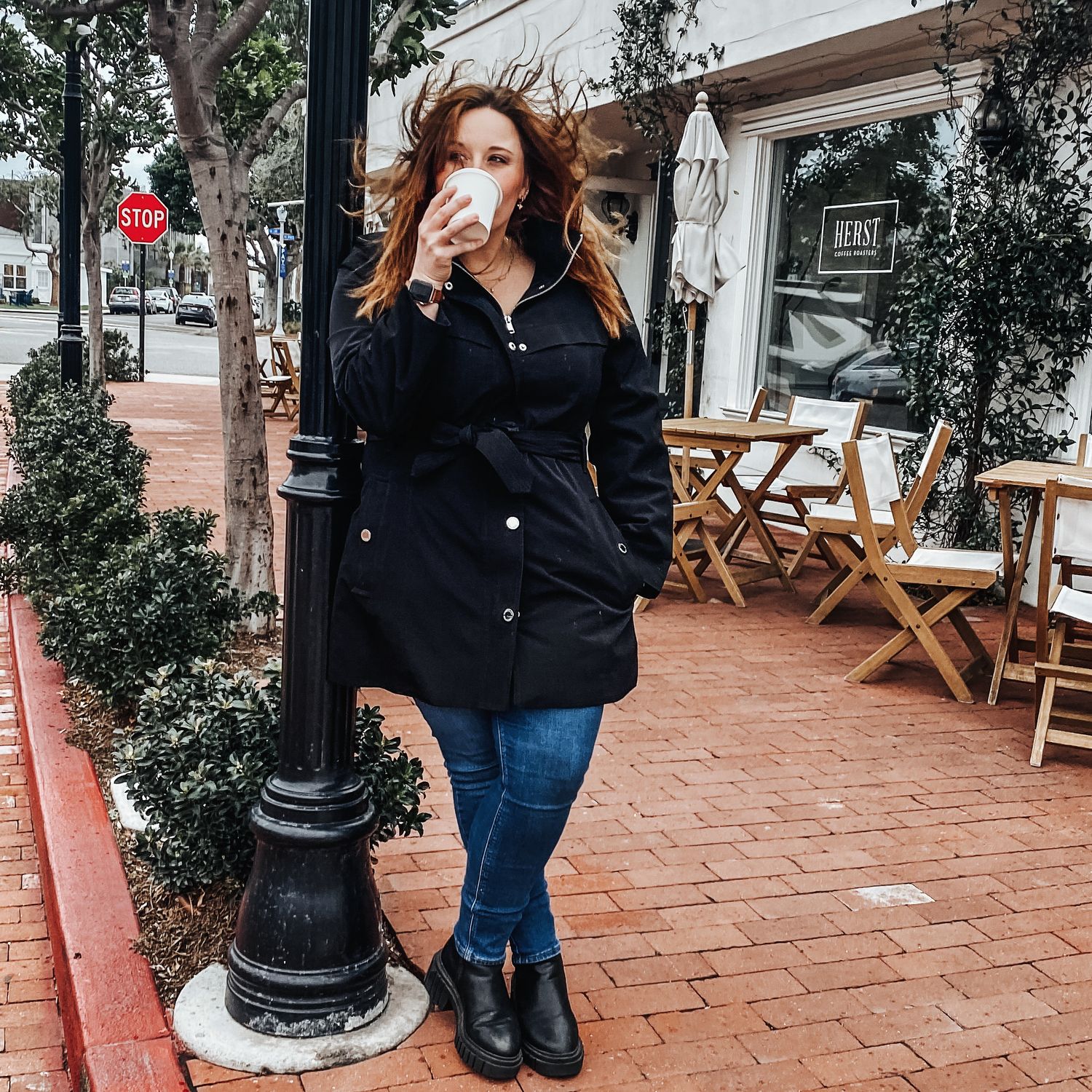 Woman in black jacket drinking coffee while standing on a brick sidewalk outside a cafe.