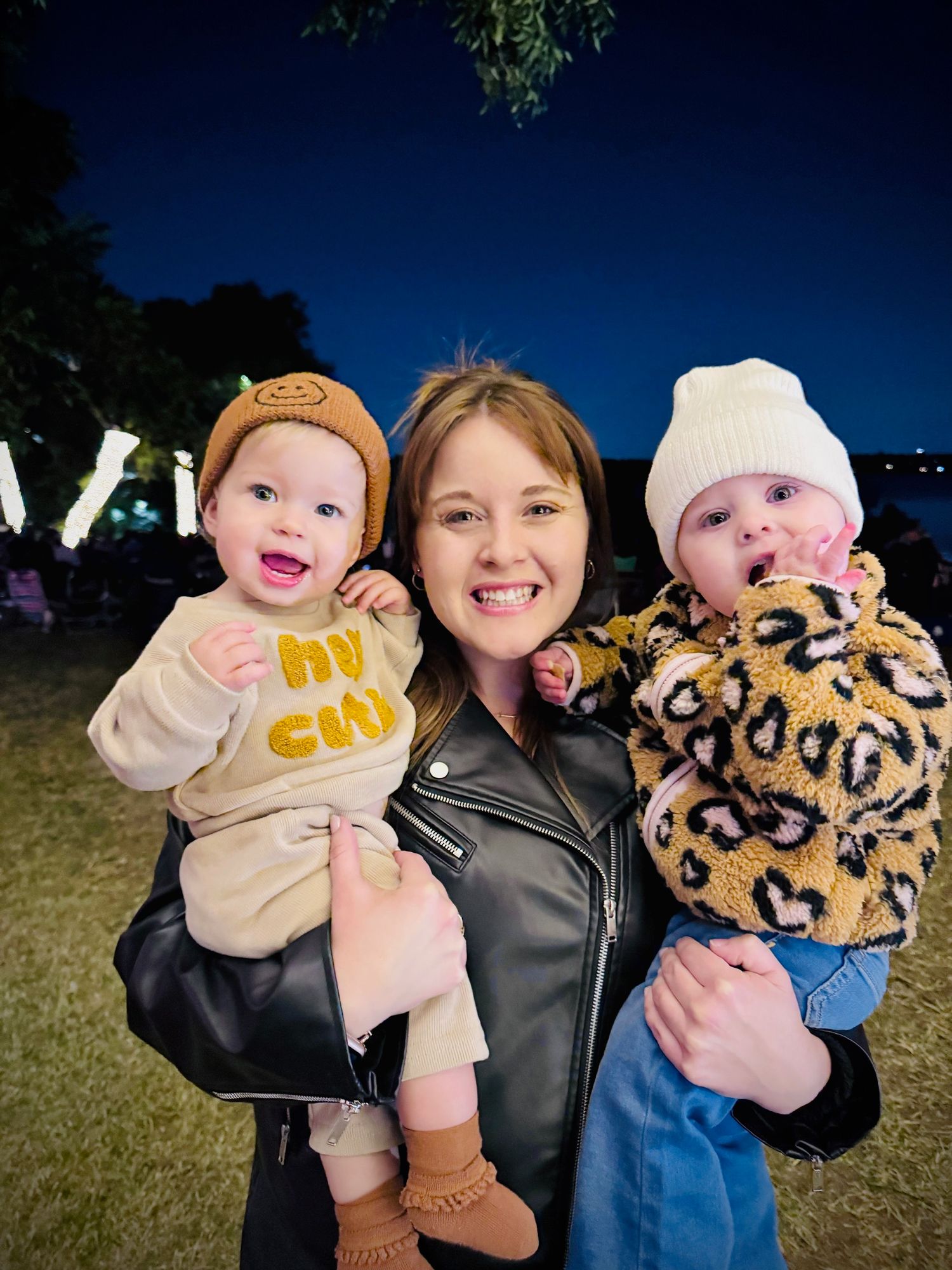 Smiling woman holding two babies in winter hats at outdoor night event with lights.