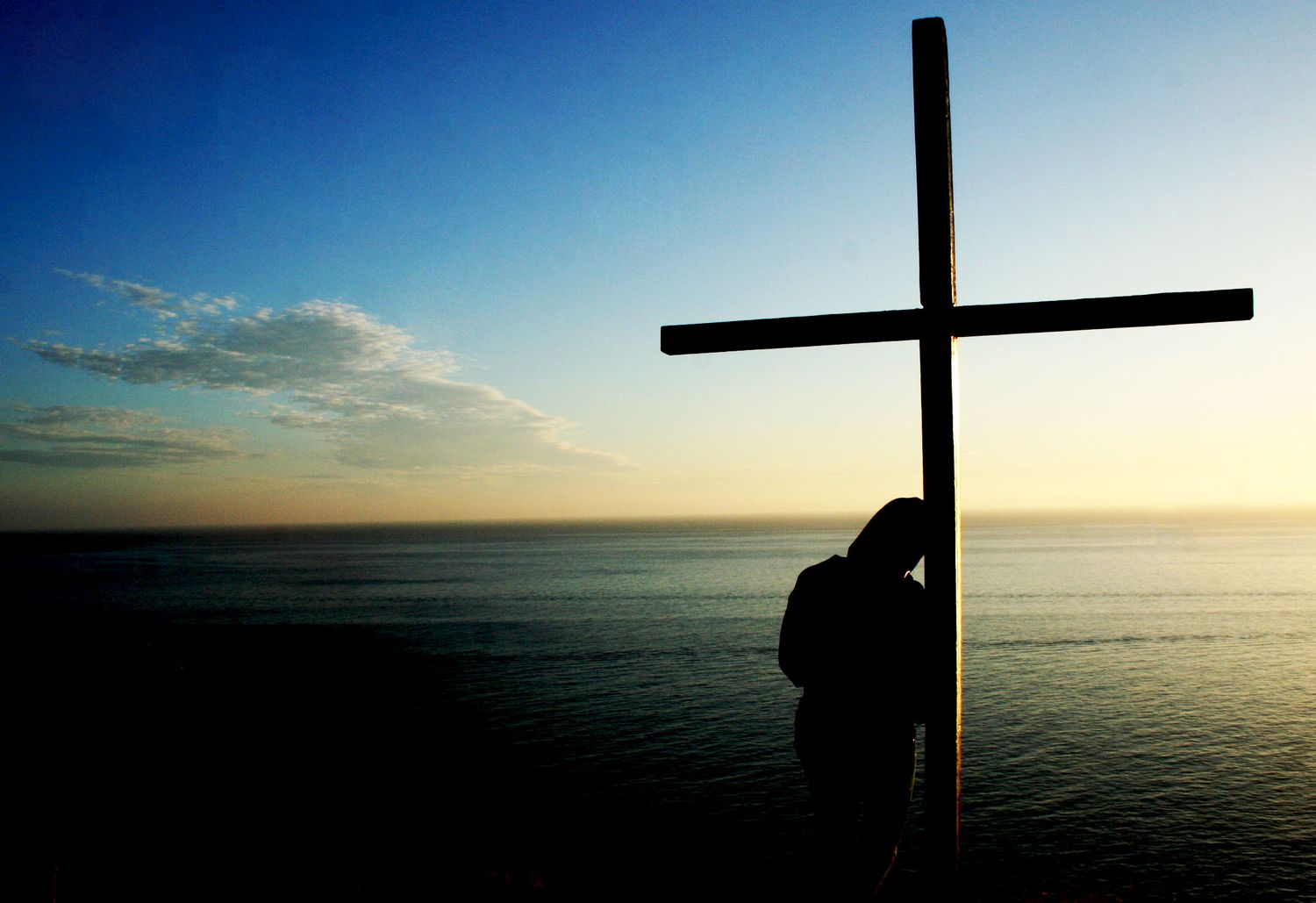 Silhouette of person leaning against wooden cross at sunset over calm ocean horizon.