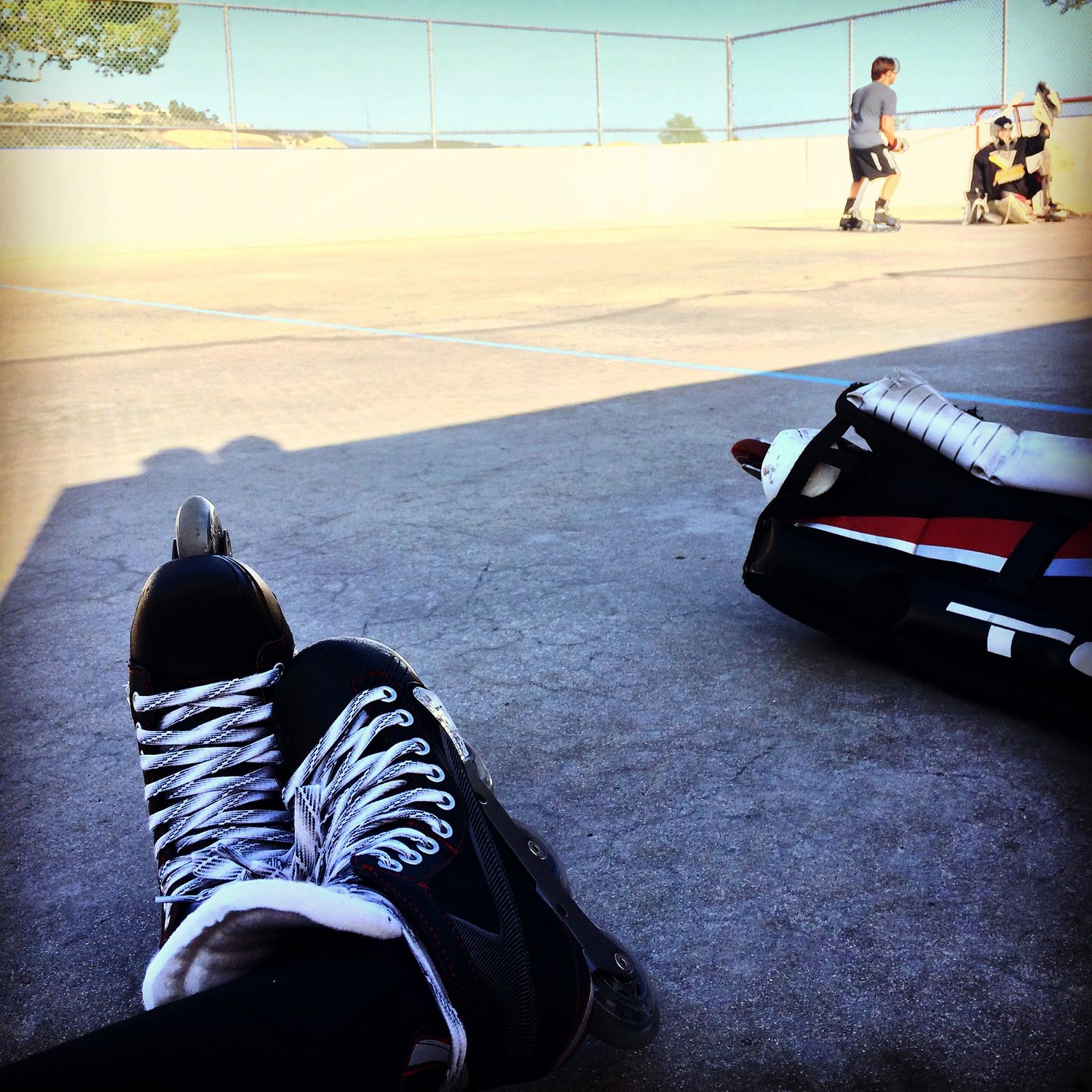 Hockey skates resting on pavement with players and sports bag in background at outdoor rink.