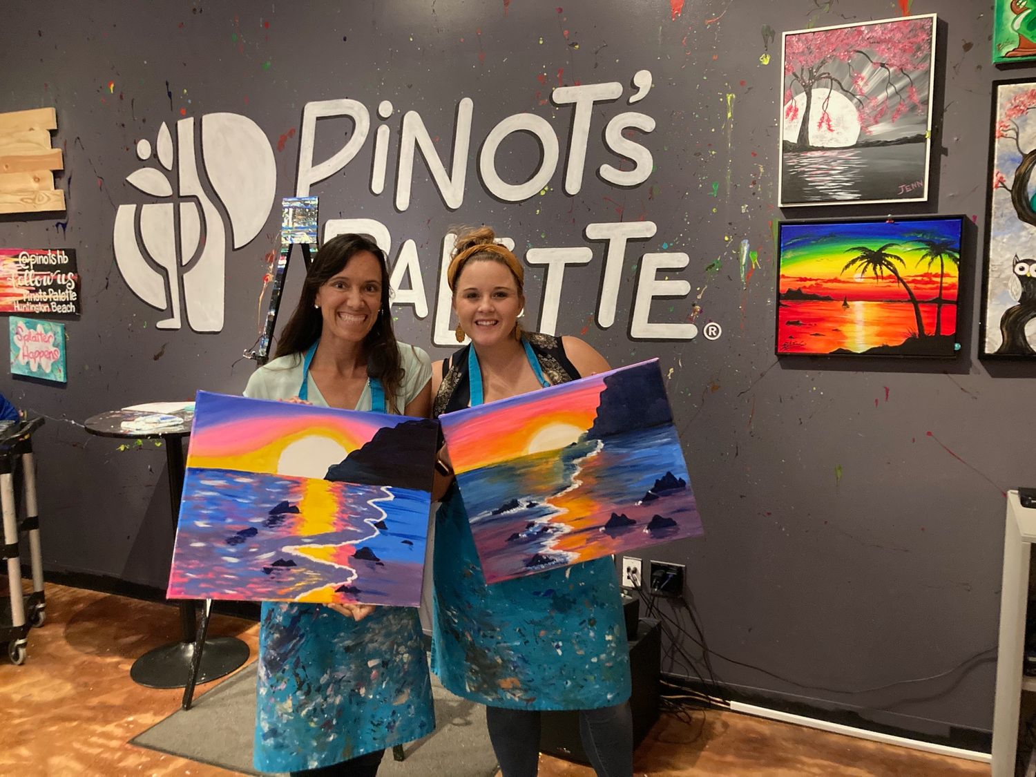Two women displaying colorful sunset paintings at Pinots Palette painting class studio.