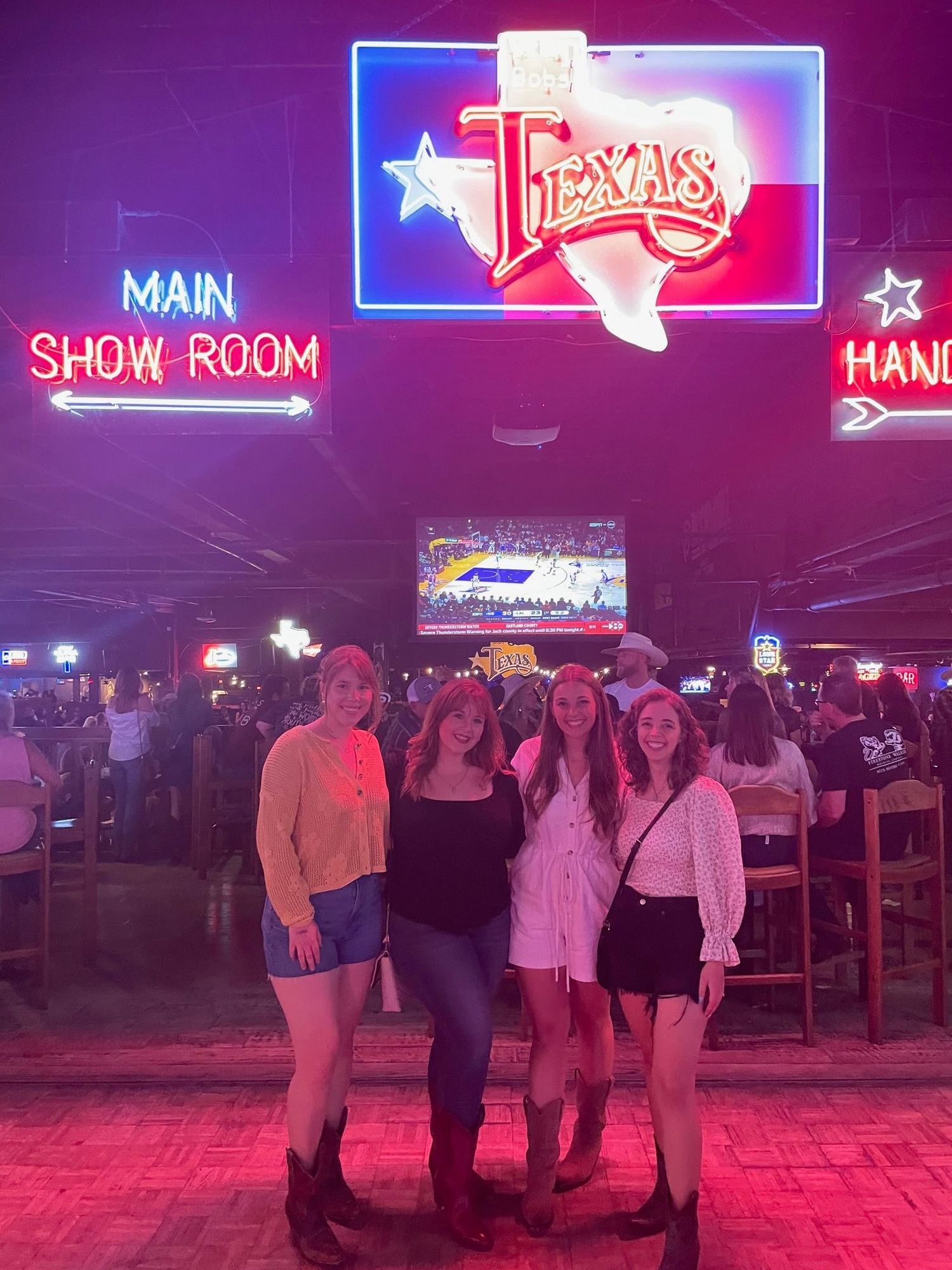 Group of five women posing under neon Texas signs in lively country western bar.