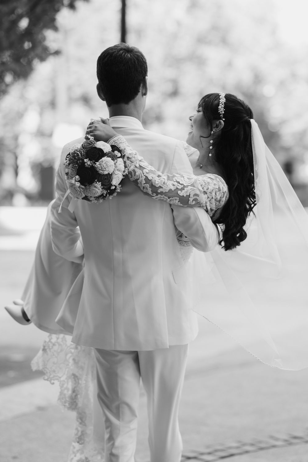 Black and white photo of a bride embracing groom from behind while holding a floral bouquet outdoors.