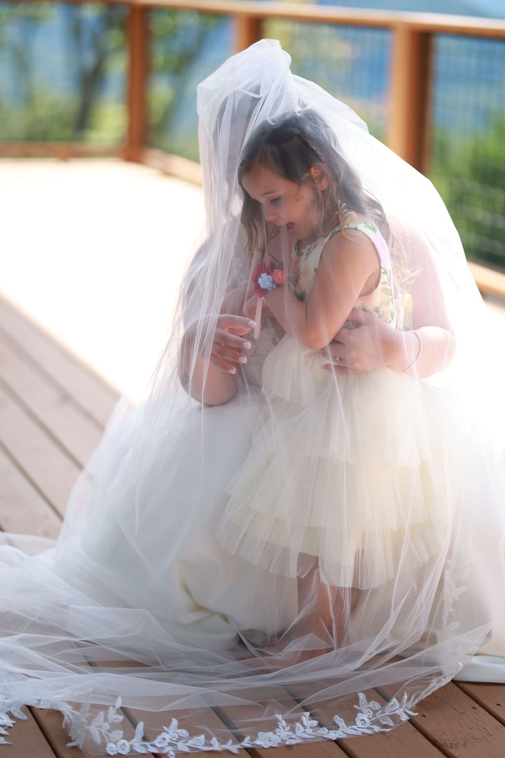 Young flower girl peeking under bride's white lace wedding veil on wooden deck during wedding ceremony.
