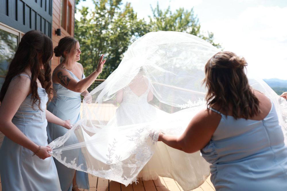 Bridesmaids in blue dresses helping bride with flowing white lace veil outdoors before wedding ceremony.
