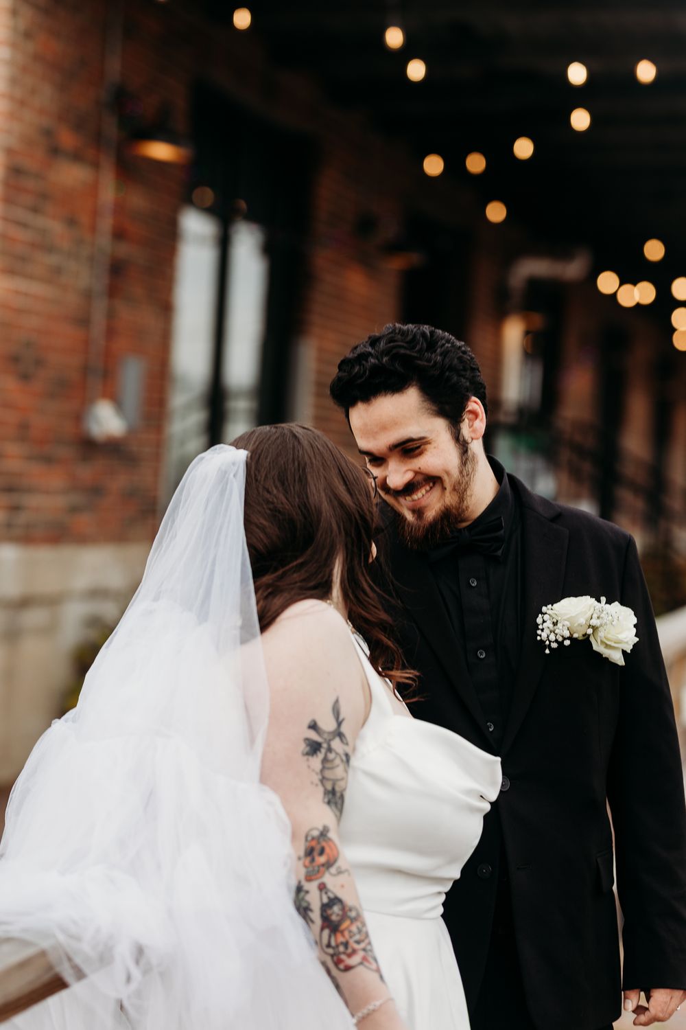 Groom in black suit smiles at tattooed bride in white gown under string lights outdoors.