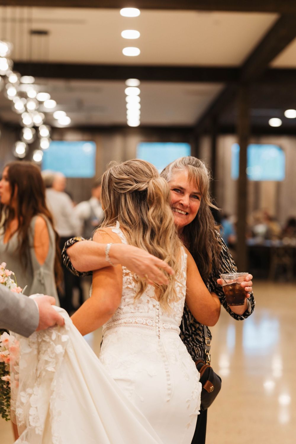 A smiling woman hugs the bride in a lace gown while holding a drink at a wedding reception.