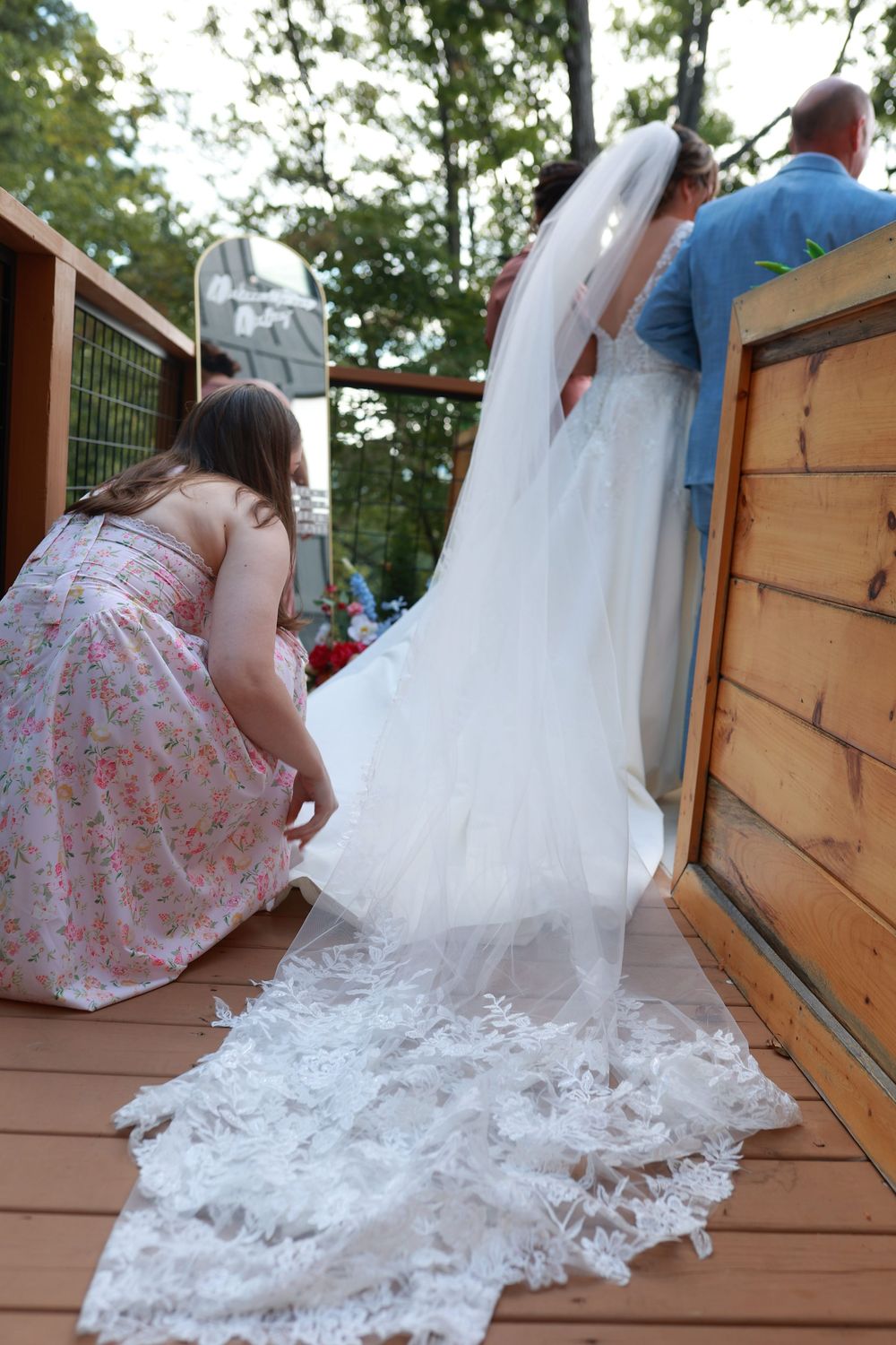 Wedding attendant arranging bride's long lace cathedral train on wooden deck during outdoor wedding preparation.