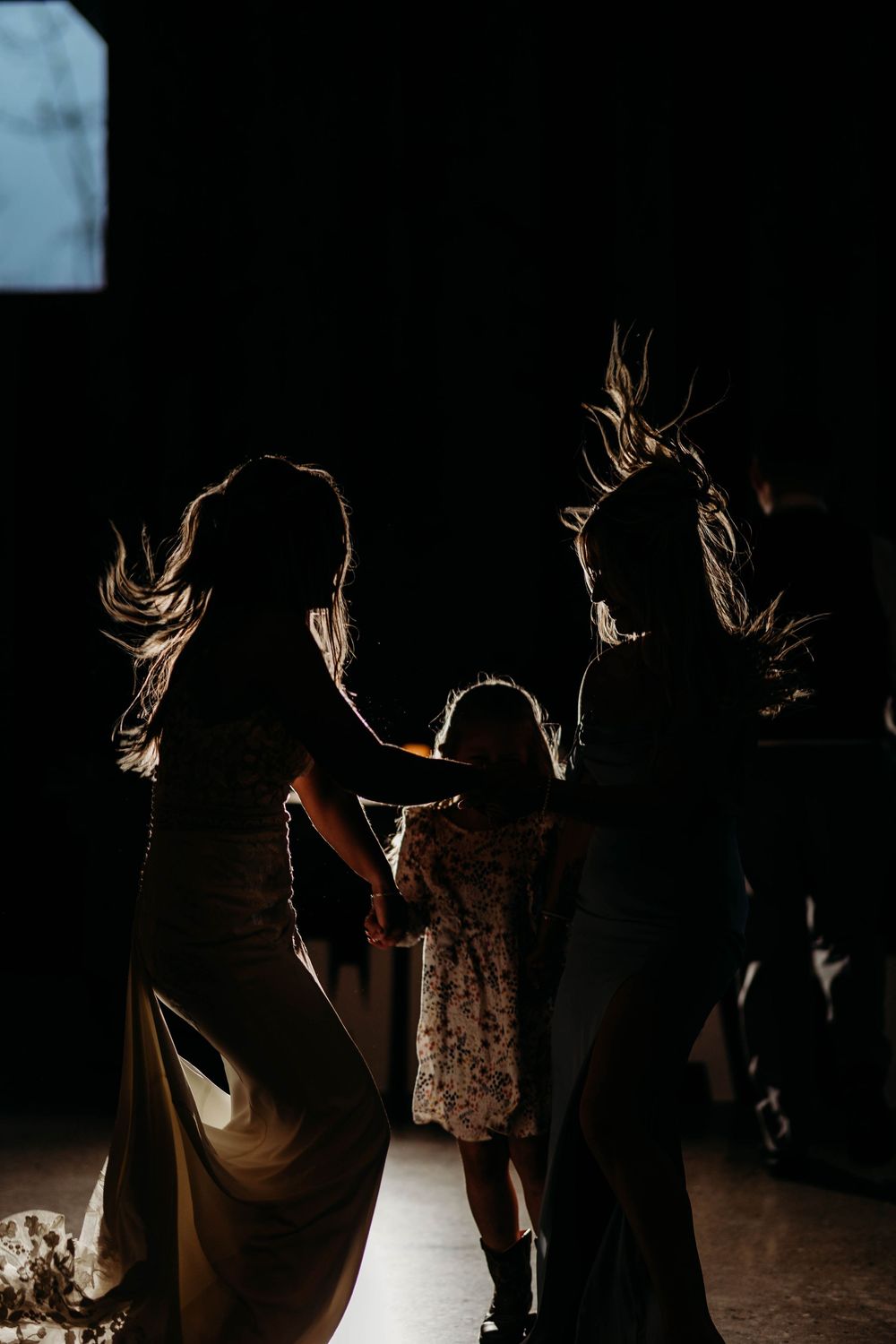 Silhouette of bride, bridesmaid, and flower girl dancing together in dramatic backlit wedding reception photo.