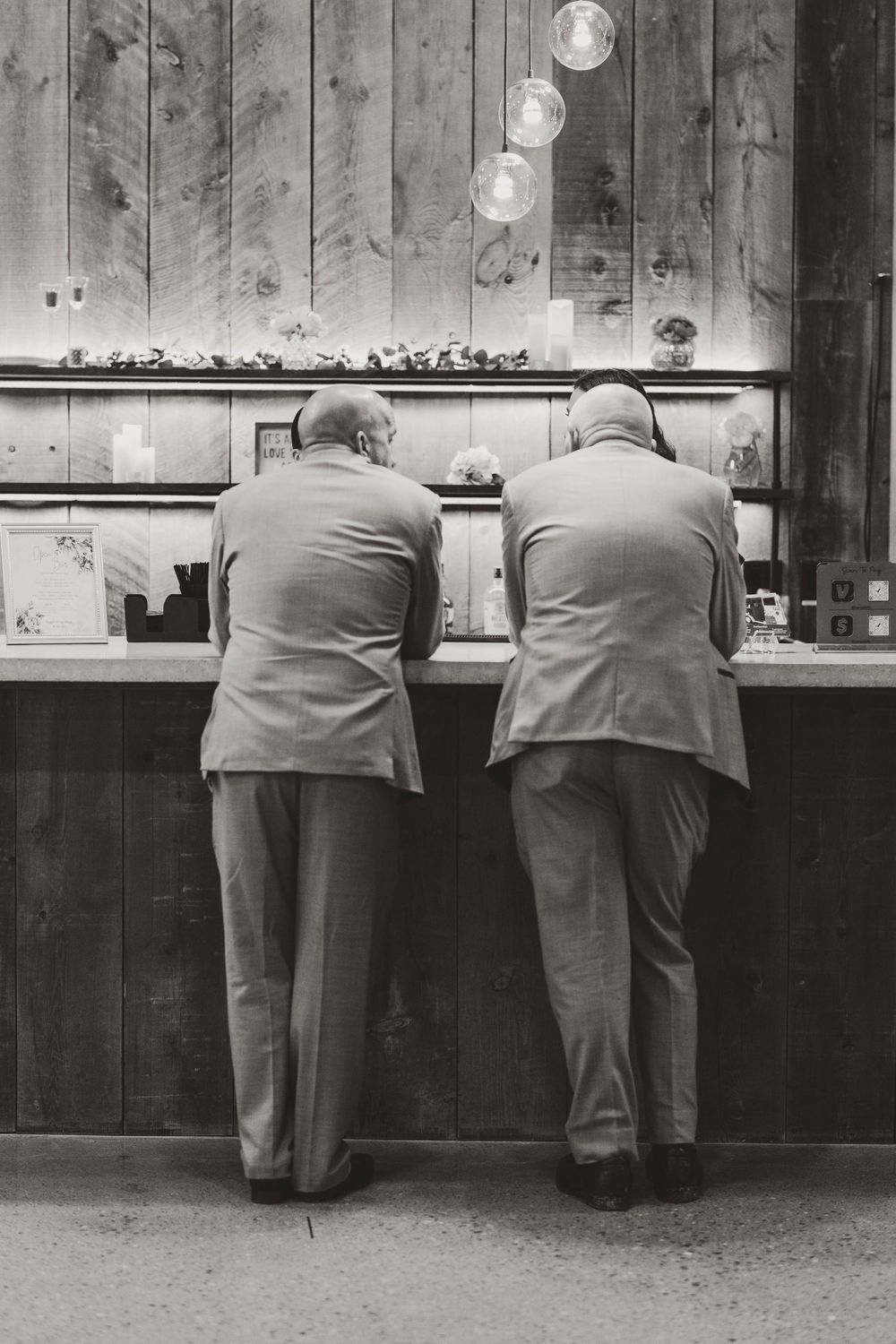 Two elderly men in suits leaning over a rustic wooden display counter at a wedding venue.