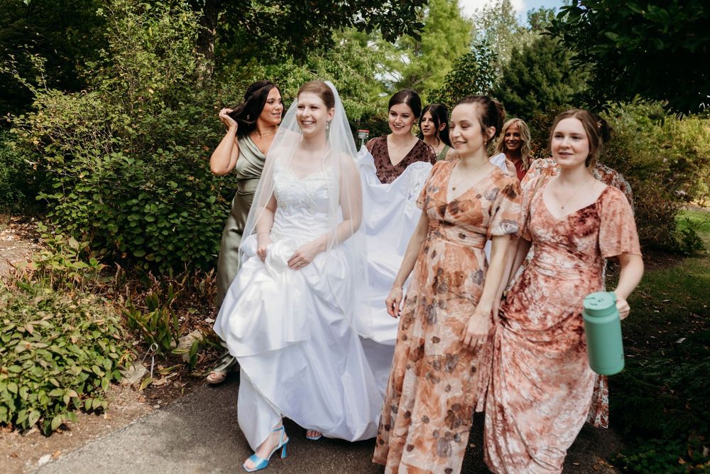 Bride in white gown walks with bridesmaids in floral dresses through a lush garden path.