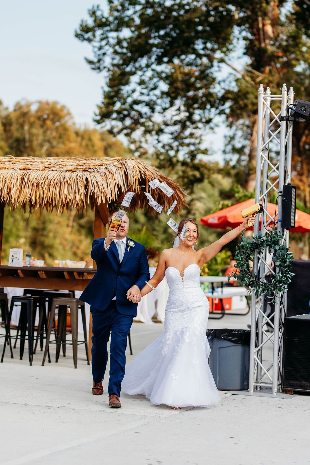 Bride in white lace gown and groom in navy suit make a grand entrance holding drinks at outdoor wedding reception.